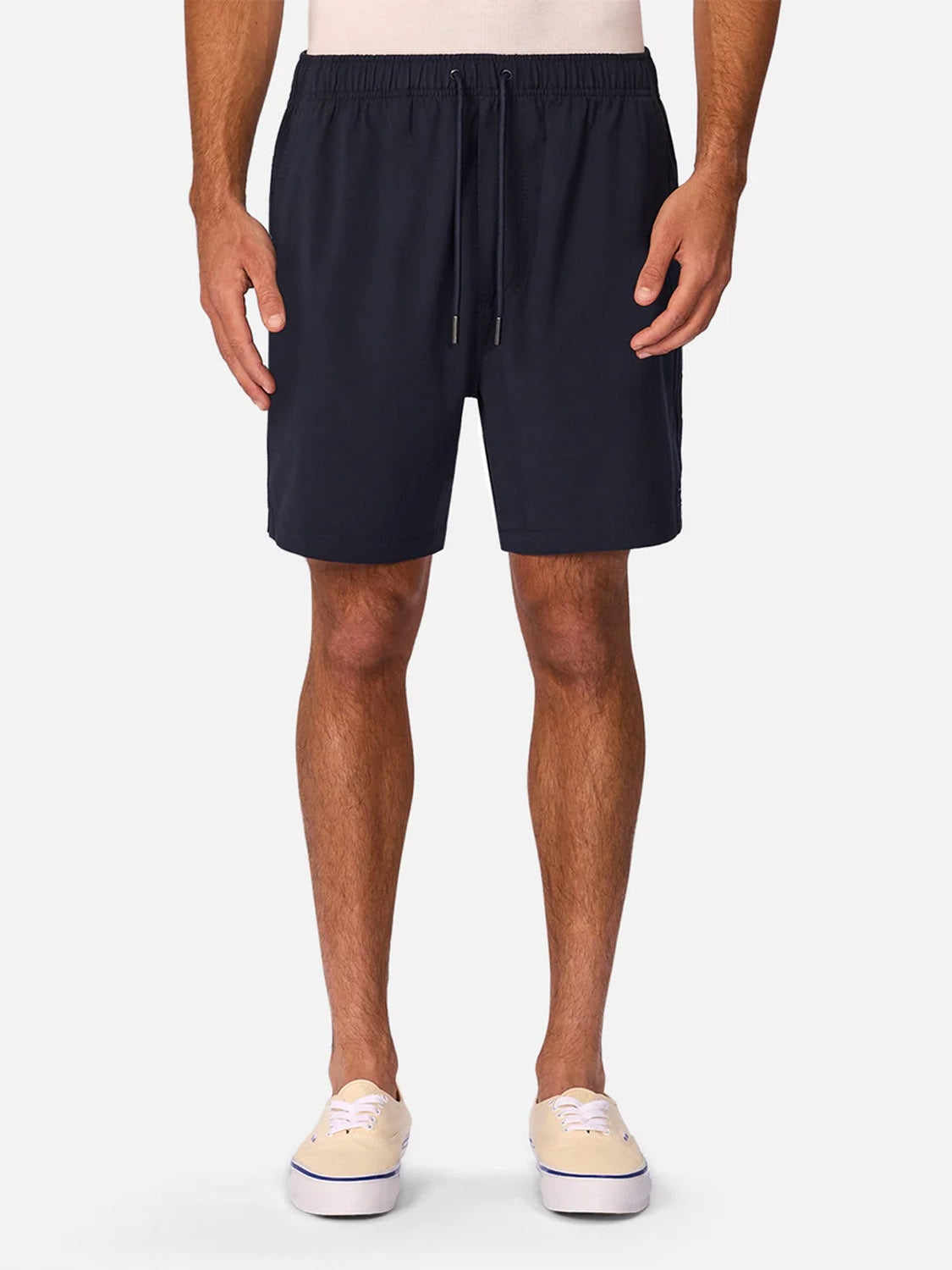 A person wearing Industrie’s The Barbados Swim Shorts in navy blue with an elastic waistband, a light-colored shirt, and beige slip-on shoes, stands against a plain white background.
