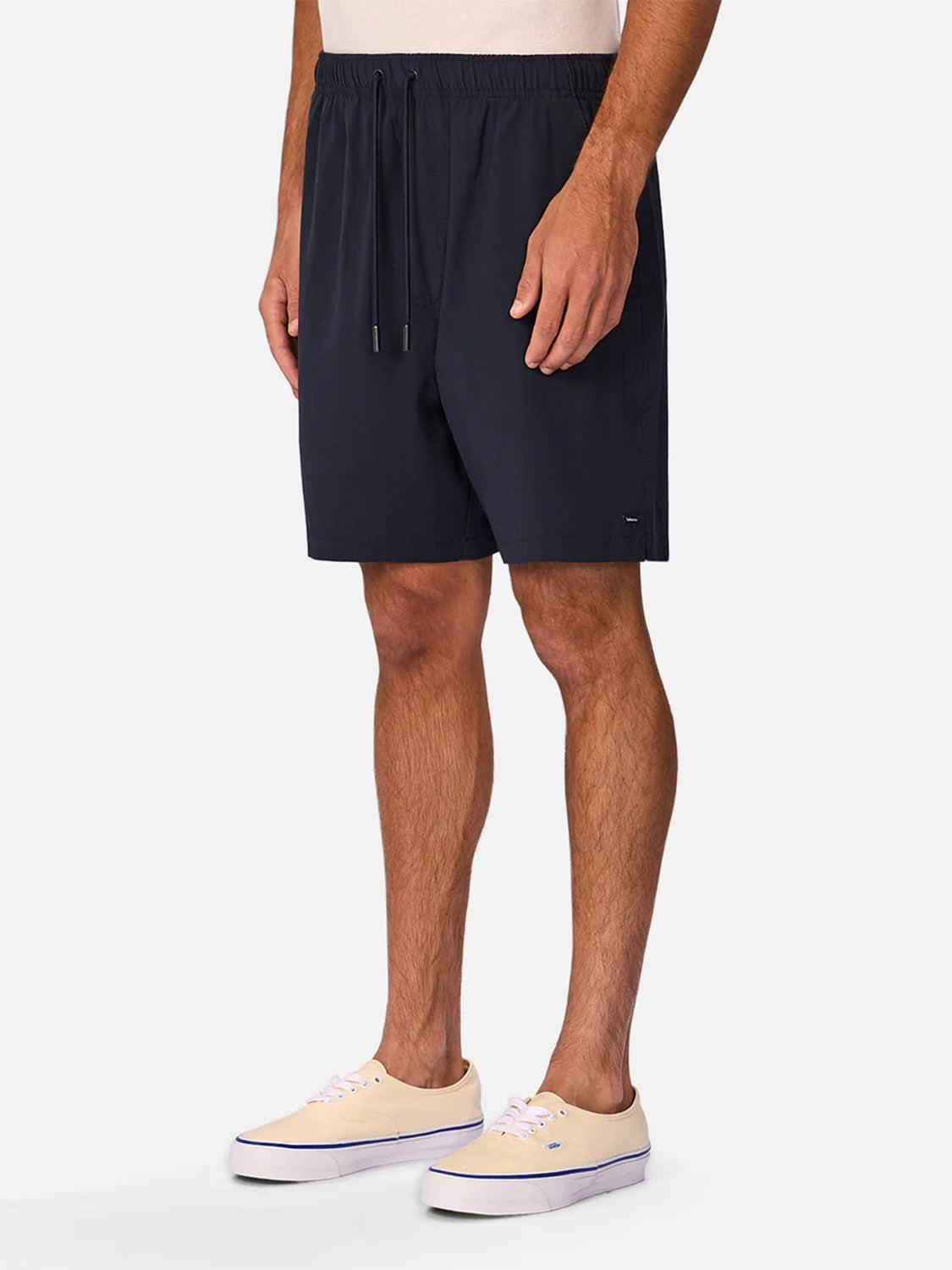A person wearing Industrie’s The Barbados Swim Shorts in navy blue with an elastic waistband and beige canvas sneakers stands against a plain white background, showing only the lower torso, legs, and part of the arms.