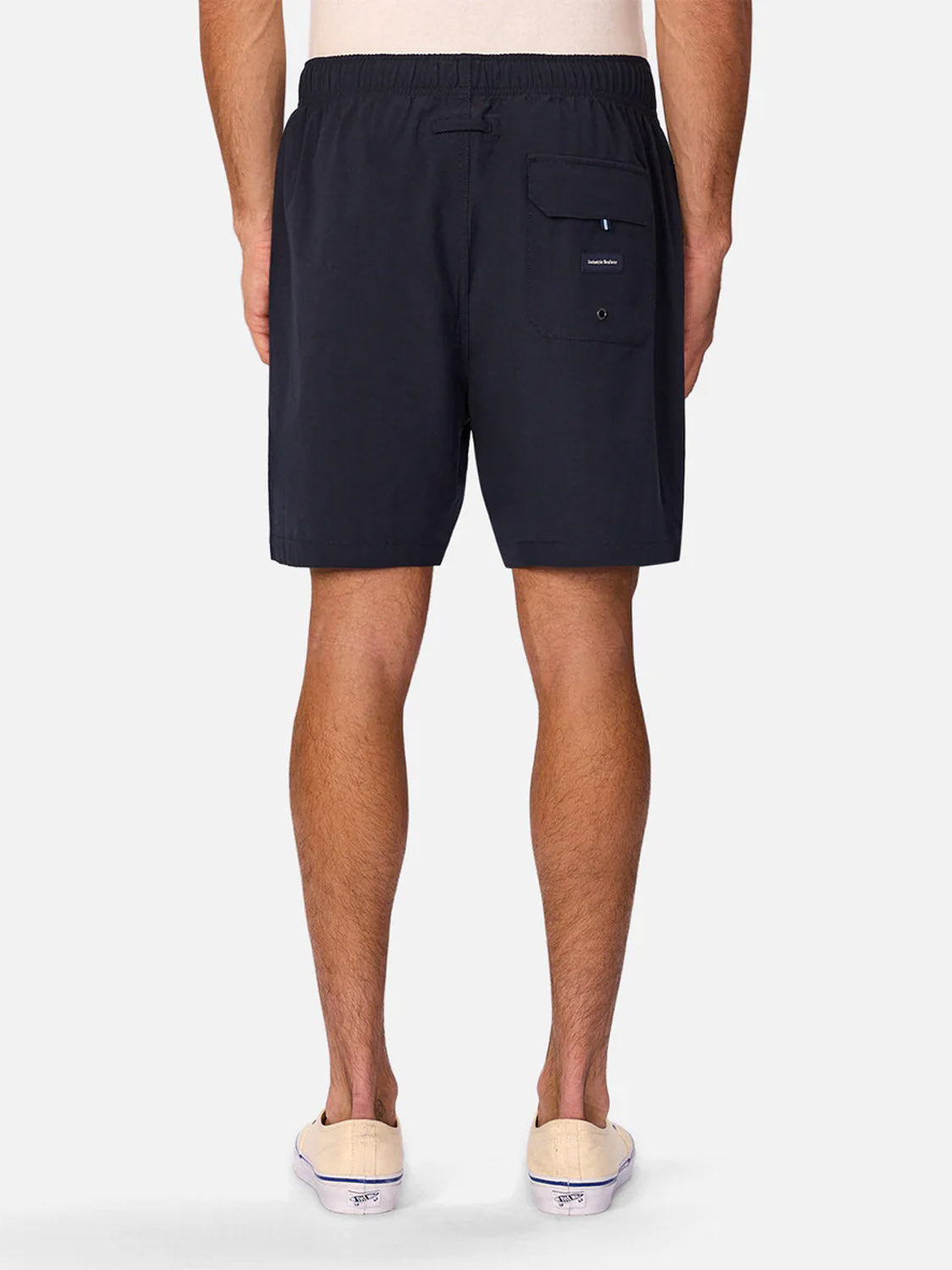 A person faces away from the camera, wearing Industrie’s The Barbados Swim Shorts in black with a back pocket and elastic waistband, paired with white slip-on shoes against a plain white background.