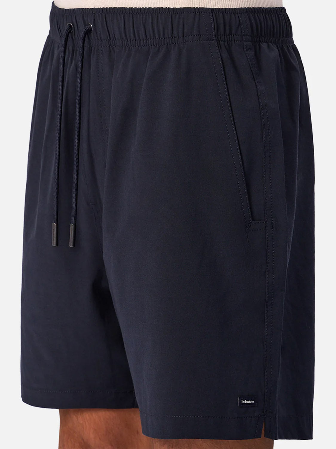A person wearing The Barbados Swim Shorts by Industrie, in dark navy with quick-dry fabric, elastic waistband, side pockets, and a small black label near the left hem, stands against a plain background.