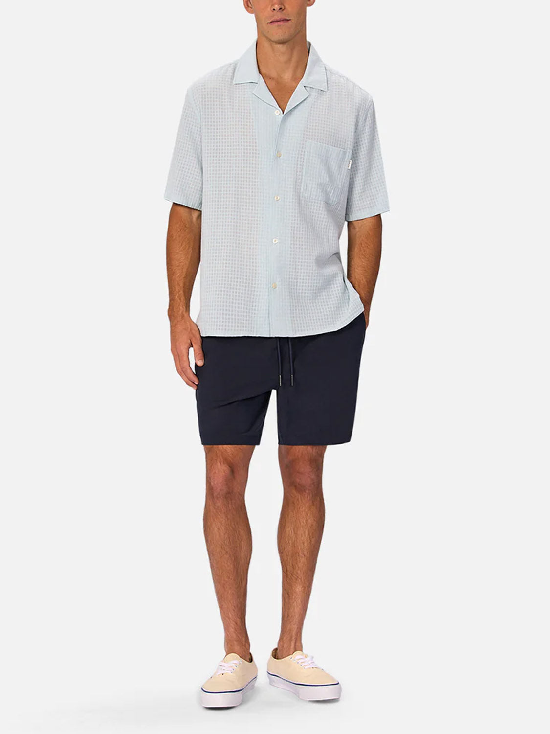 A man stands against a plain background wearing Industrie’s The Barbados Swim Shorts, featuring an elastic waistband, paired with a light short-sleeve button-up shirt and light sneakers. His left hand is in his pocket, and his right arm rests at his side.