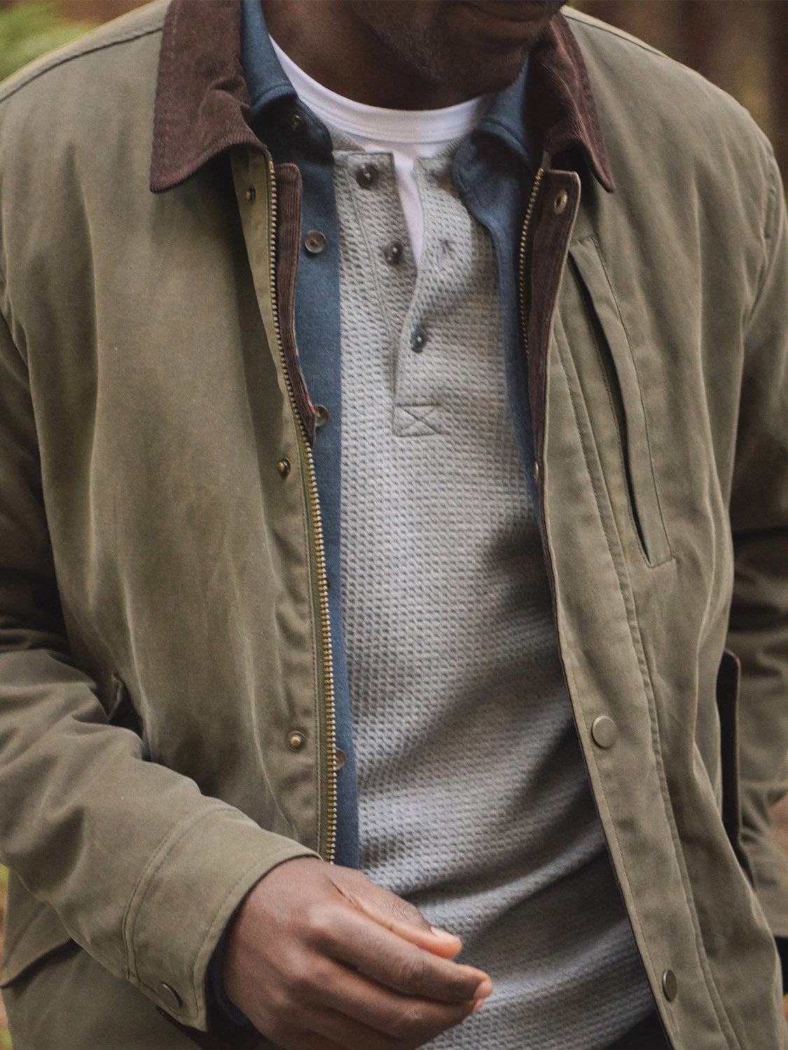 A person wearing the Marine Layer Weston Wax Canvas Barn Jacket over a blue and gray collared shirt stands outdoors, partially out of frame, holding something in their right hand.