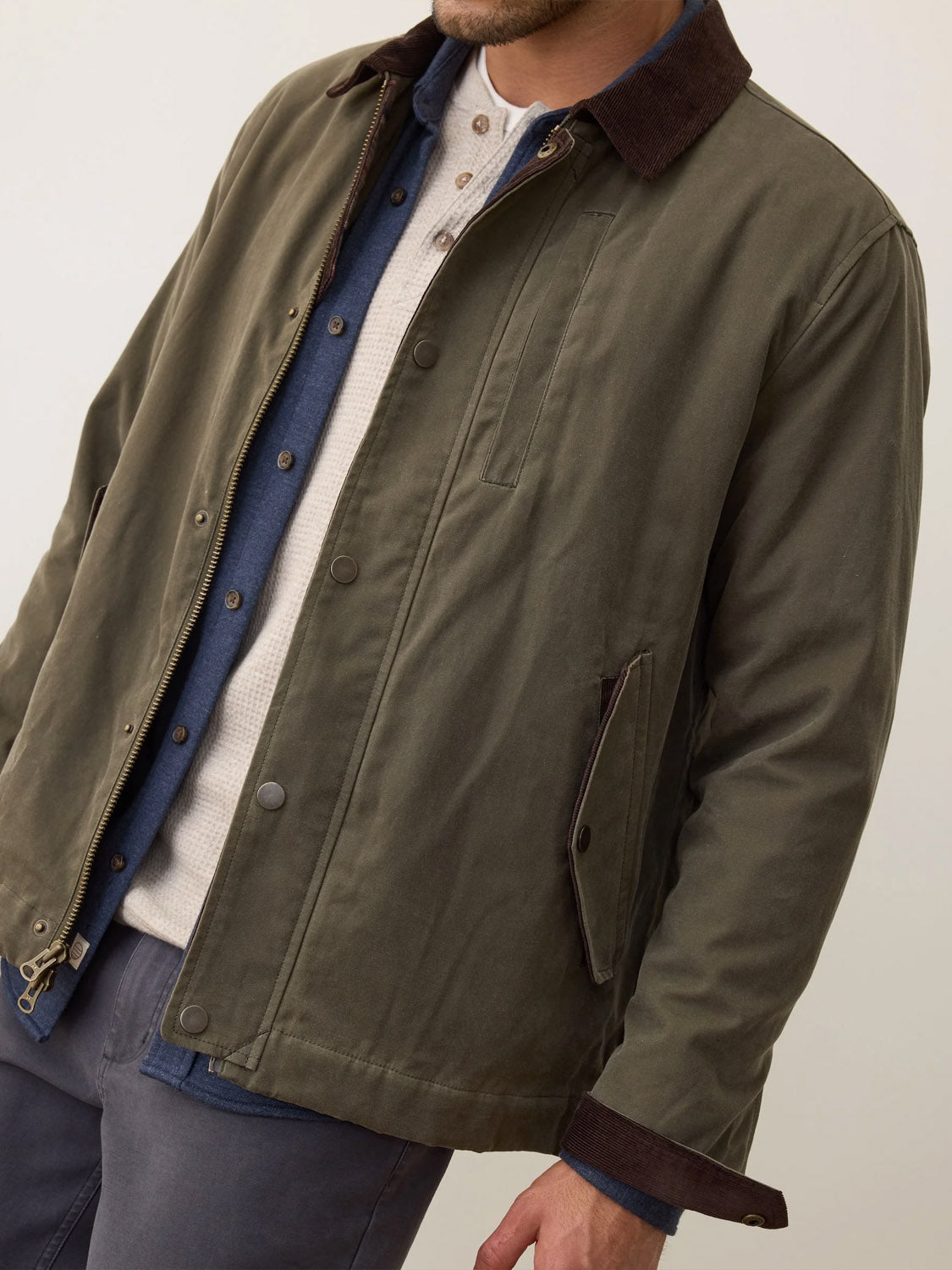 A man wears the Marine Layer Weston Wax Canvas Barn Jacket in olive green with a dark brown collar, layered over a blue shirt and light beige henley, paired with dark pants. The cropped image shows from shoulders to mid-thigh.