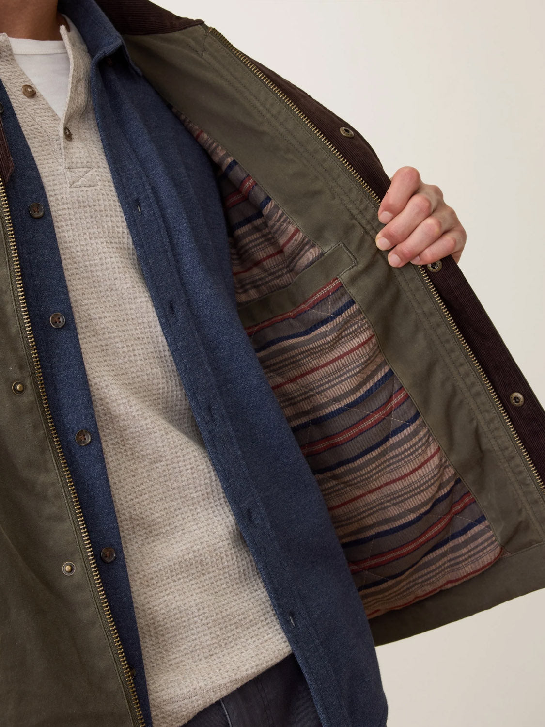 A person wears the Marine Layer Weston Wax Canvas Barn Jacket open, showing its striped red, grey, and blue lining, layered over a white textured shirt and blue button-down. Only their upper body is visible.