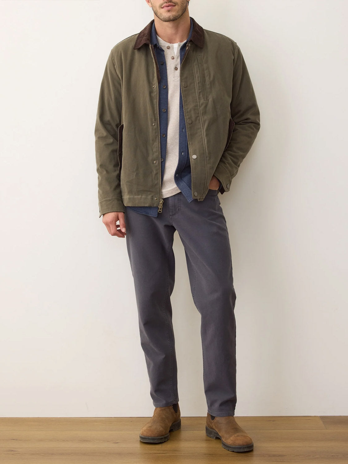 A man (face not shown) stands against a white wall wearing Marine Layer’s Weston Wax Canvas Barn Jacket, featuring a dark collar, layered over a blue shirt and beige henley, paired with gray pants and brown suede boots.