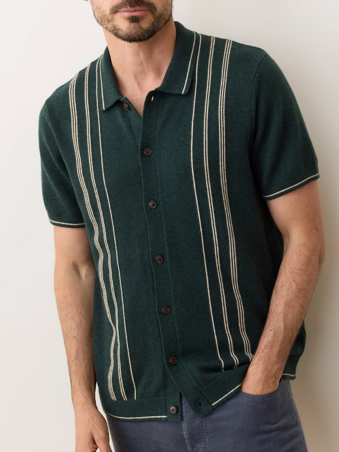 A man wears the Marine Layer Barry Sweater Button Down, a dark green short-sleeve knit shirt with cream vertical stripes and dark buttons, standing against a light background with his hands in his pockets.