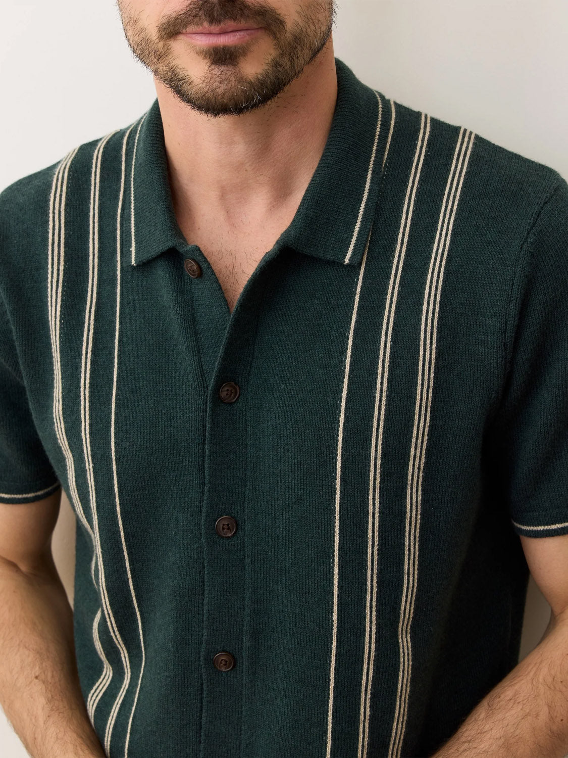 A man wearing the Marine Layer Barry Sweater Button Down, a classic fit dark green knit shirt with cream stripes and brown buttons, stands against a light background. His face is partially visible with a neat beard and mustache.