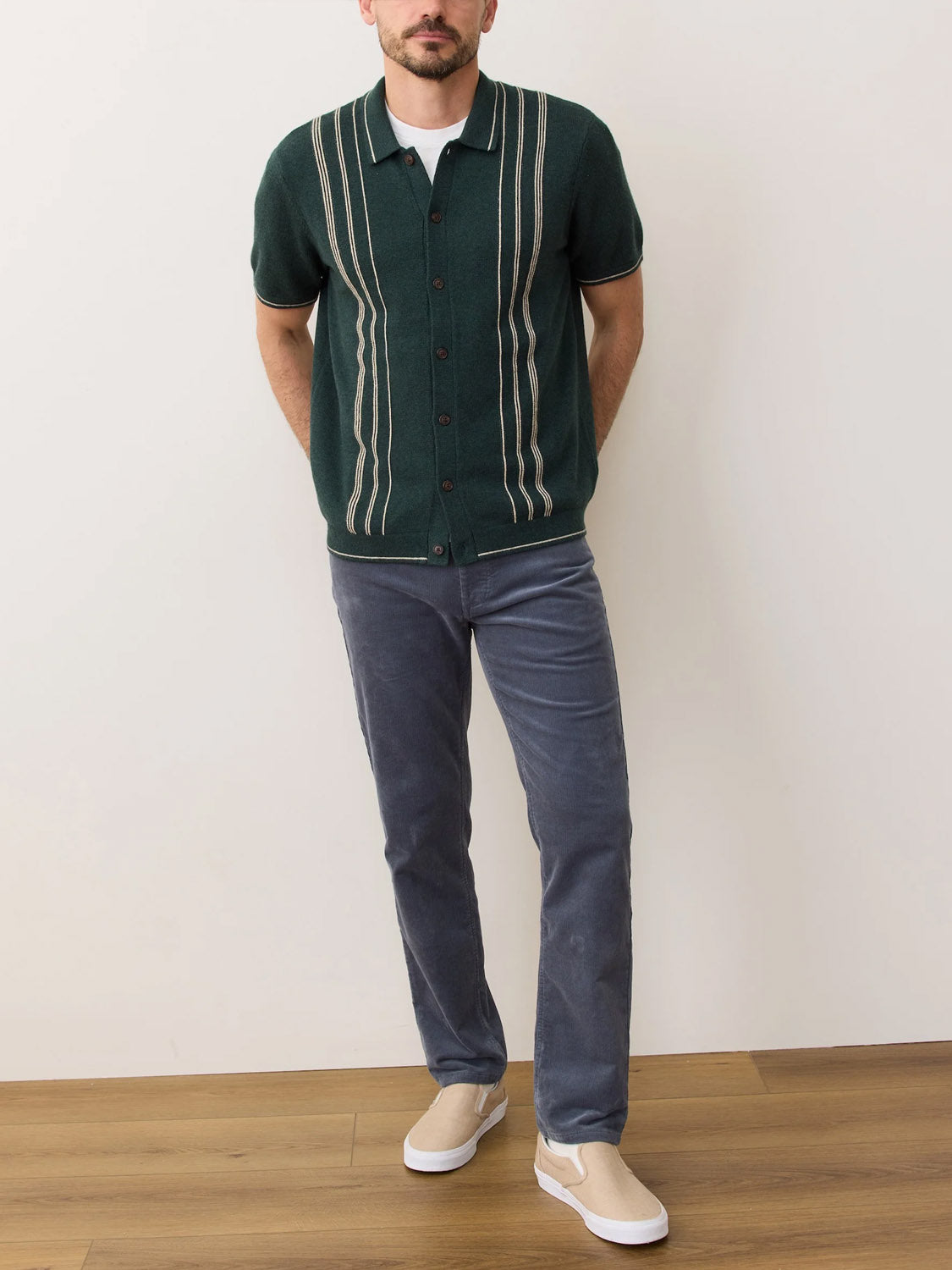 A man stands indoors against a white wall, wearing the Marine Layer Barry Sweater Button Down in dark green with white stripes, paired with gray pants and beige slip-on shoes. He has his hands behind his back.