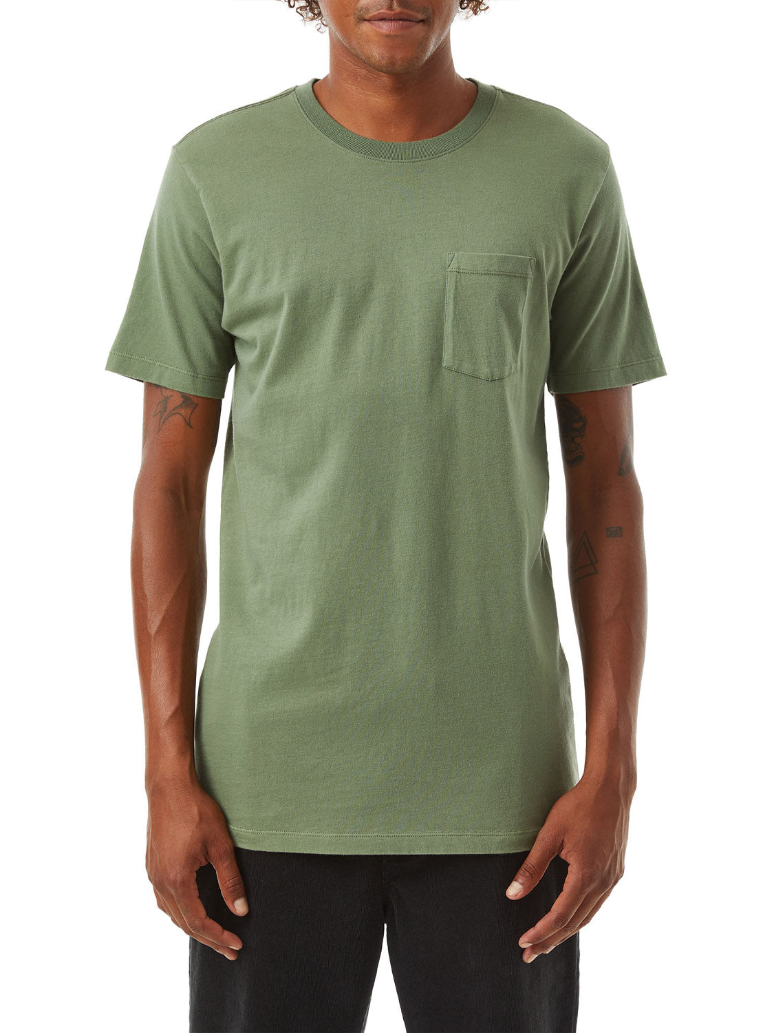 A person in a Katin Base Tee, an olive green, garment-dyed organic cotton pocket tee by Katin, stands against a white background with tattooed arms, paired with dark pants.