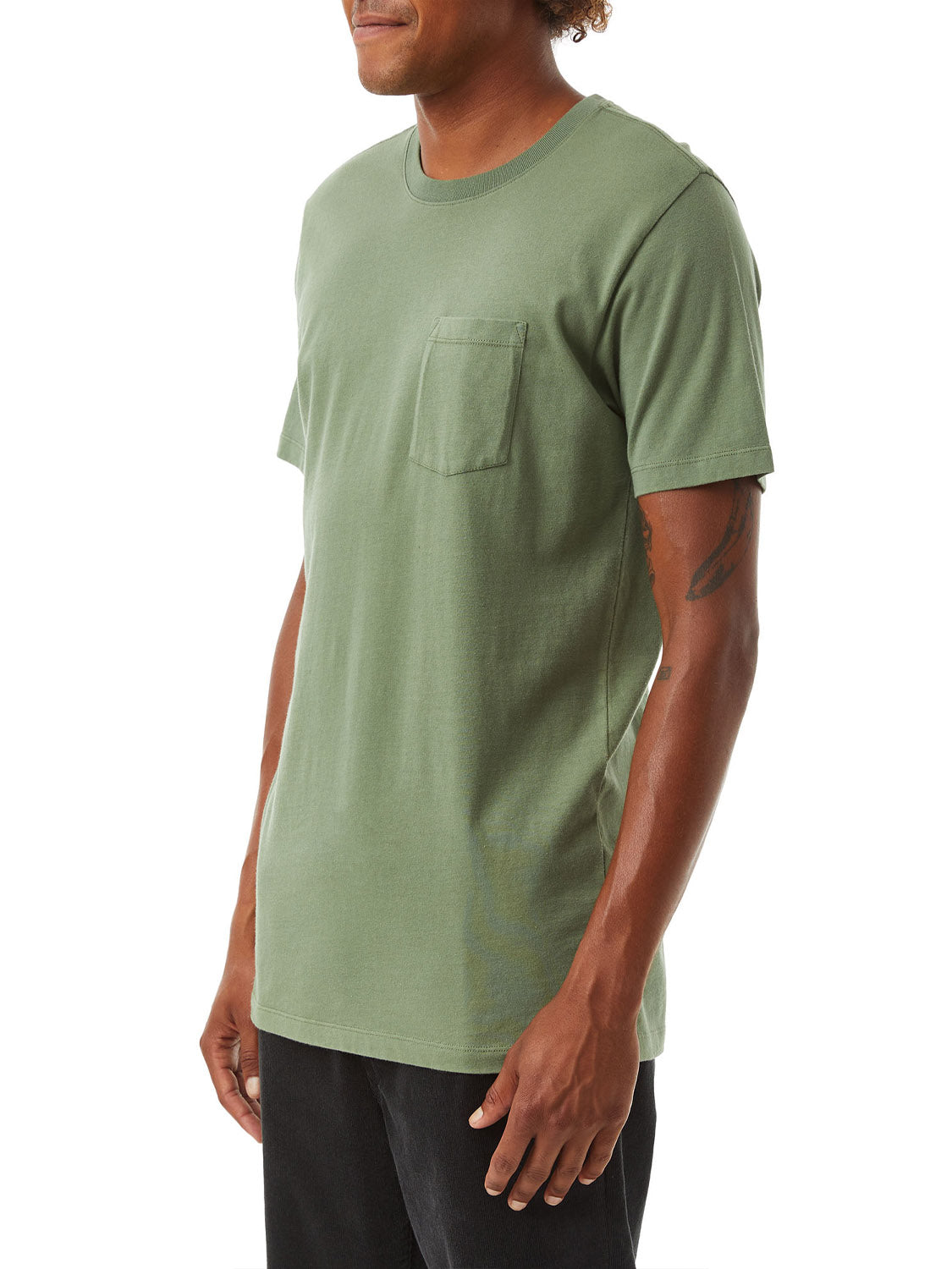 A person in a side view wears the Katin Base Tee, a plain green garment-dyed organic cotton tee by Katin, with short sleeves and a visible tattoo on their right arm. They pair it with black pants against a white background.