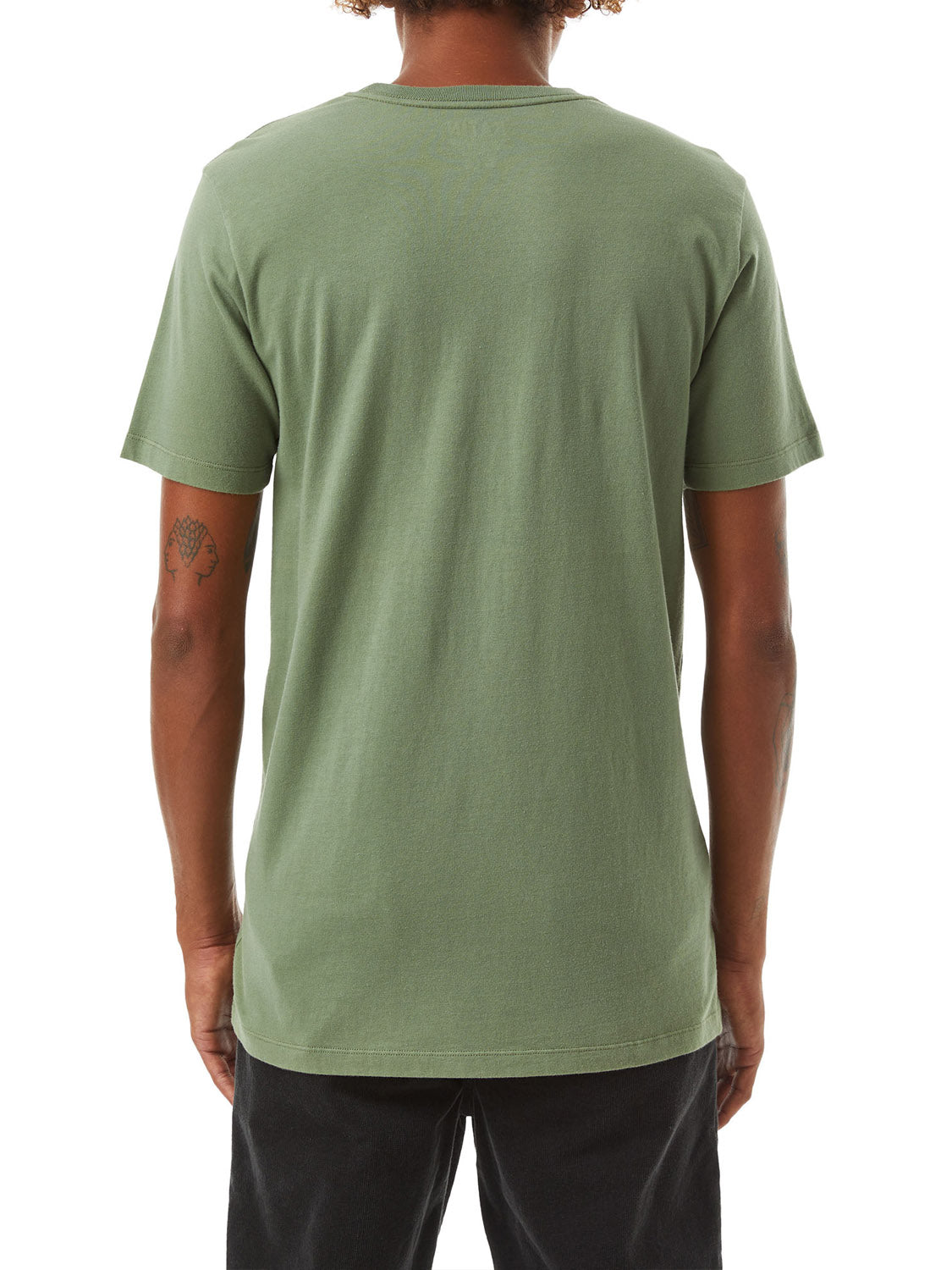 The person is wearing a Katin Base Tee in plain green, made of organic cotton, paired with black pants. Their curly hair flows down, and tattoos are visible on their left arm from the back view.