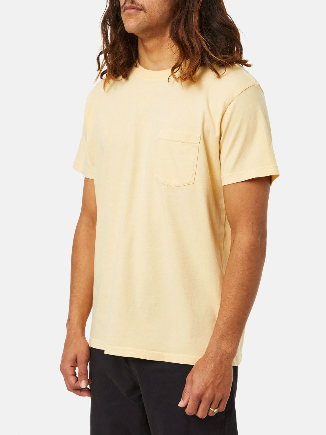 A person with long brown hair wears a Katin Base Tee in pale yellow organic cotton, paired with black pants against a plain white background.