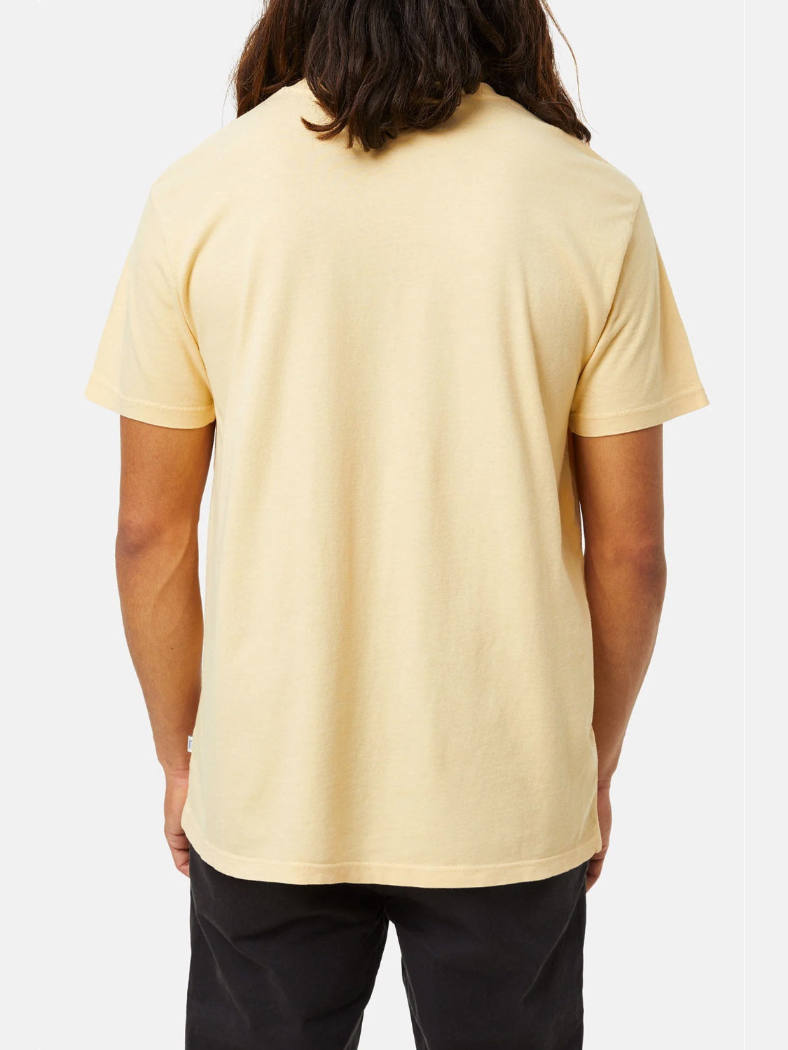 The rear view captures a person with long hair wearing the Katin Base Tee in light yellow and dark pants. Made from organic cotton, this Katin T-shirt is set against a plain white background, highlighting its soft hues and sustainable design.