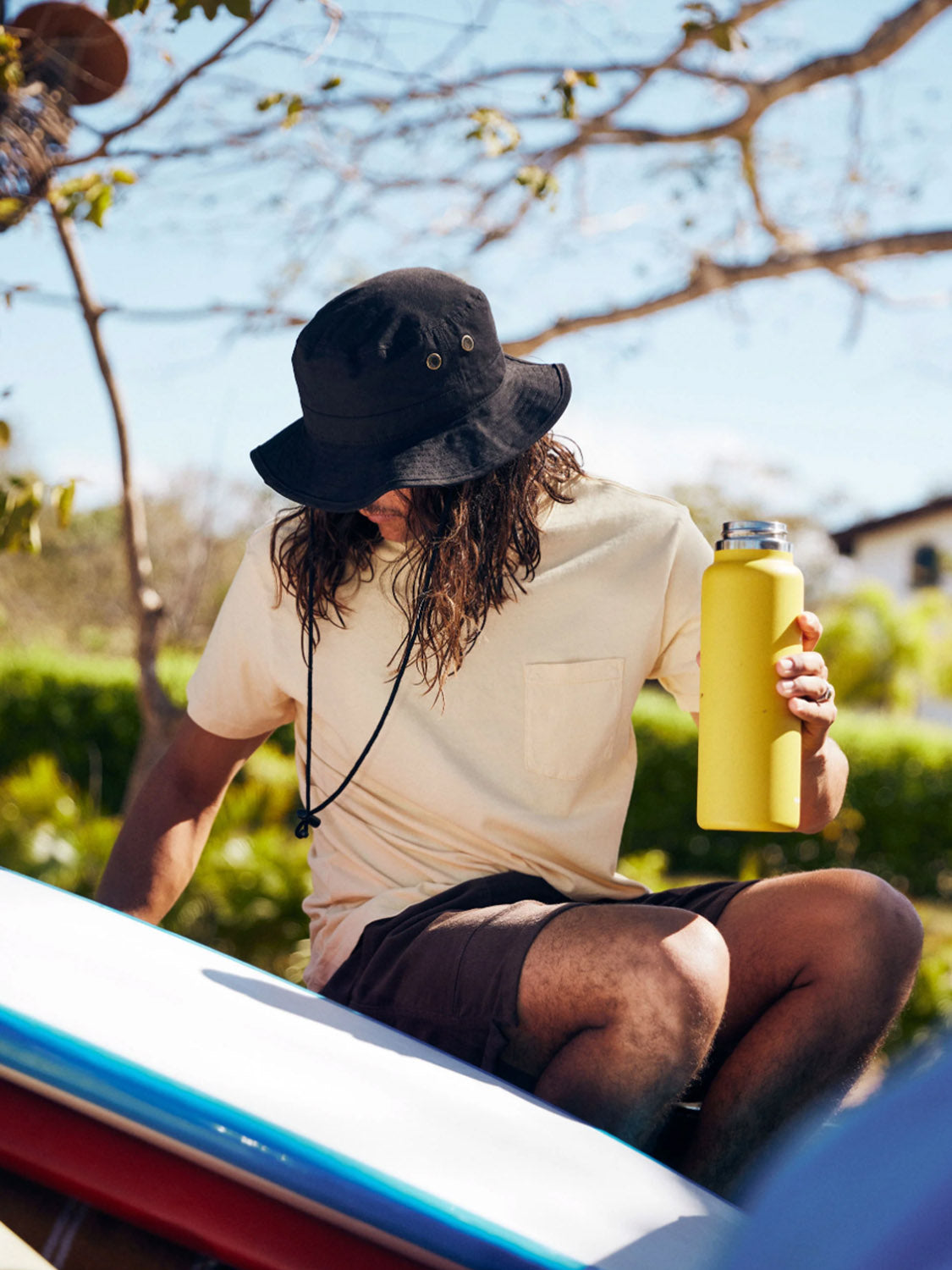 A person with long hair, wearing a black bucket hat and the Katin Base Tee, sits on a surfboard holding a yellow water bottle against a backdrop of greenery and clear, sunny skies.