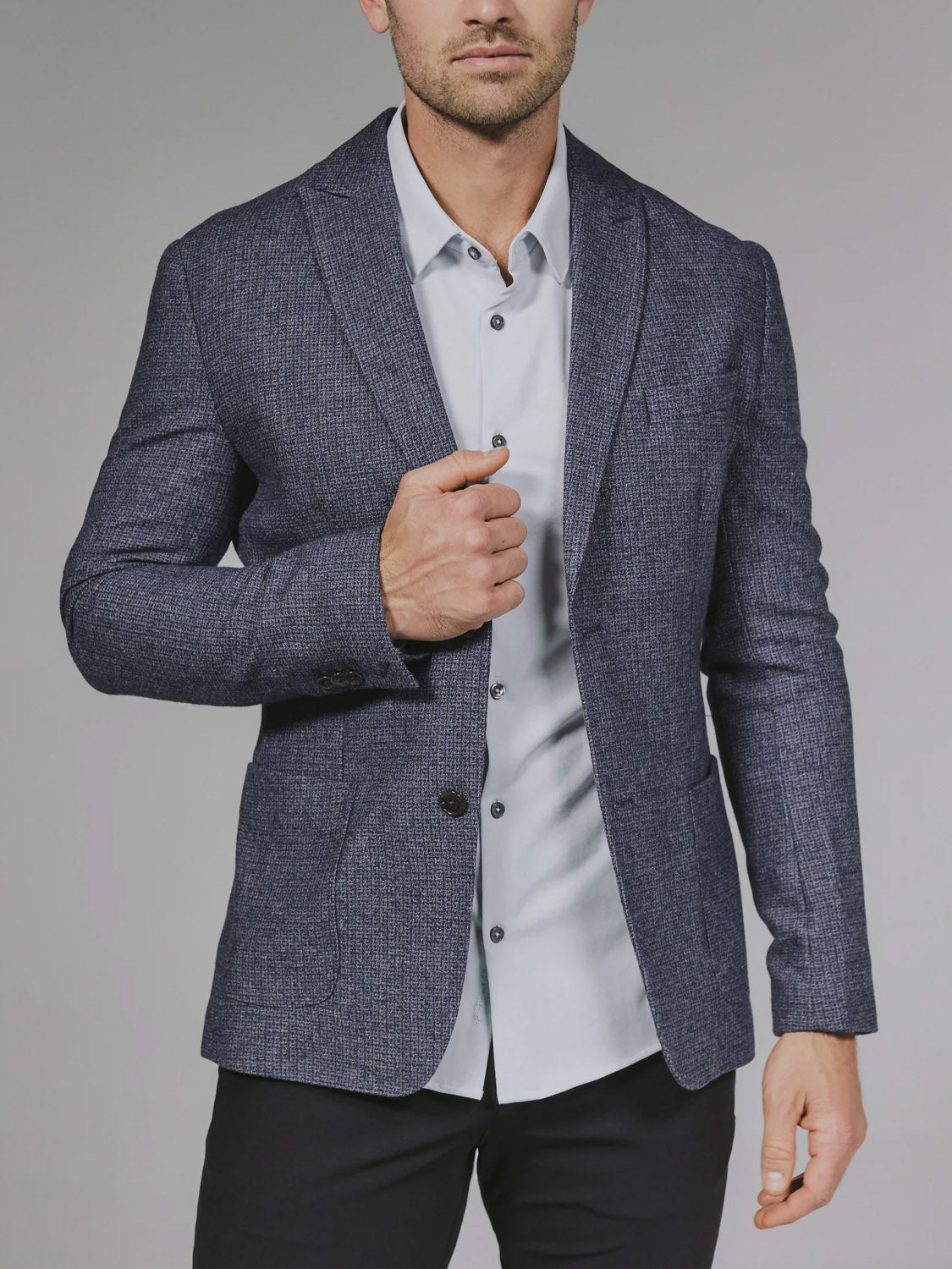A man models the 7 Diamonds Belfast Blazer in classic navy over a light gray button-up and black pants, standing against a plain gray background with his face partially out of the frame.