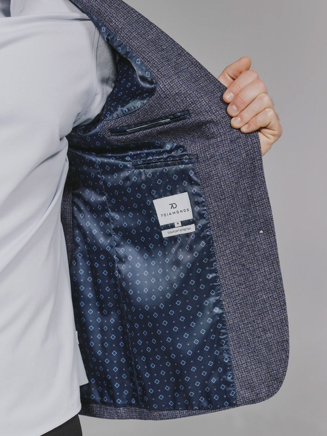 A person holds open the 7 Diamonds Belfast Blazer, revealing its blue geometric lining and white 7DIAMONDS label, while wearing a white shirt, showcasing a modern update to this classic piece.
