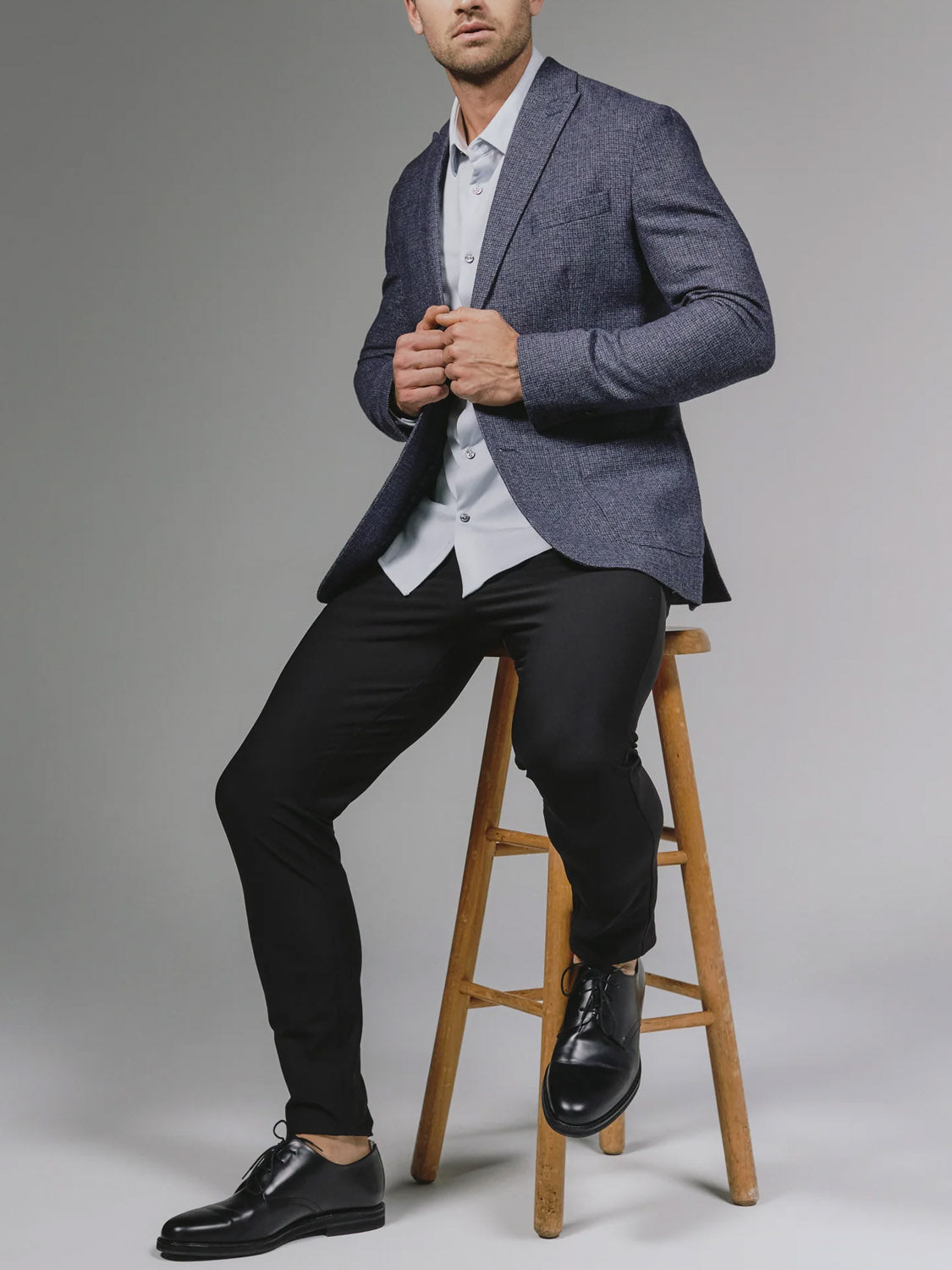 A man in the 7 Diamonds Belfast Blazer sits on a wooden stool, paired with a white shirt, black pants, and black dress shoes against a plain gray background. His face is partially out of frame.