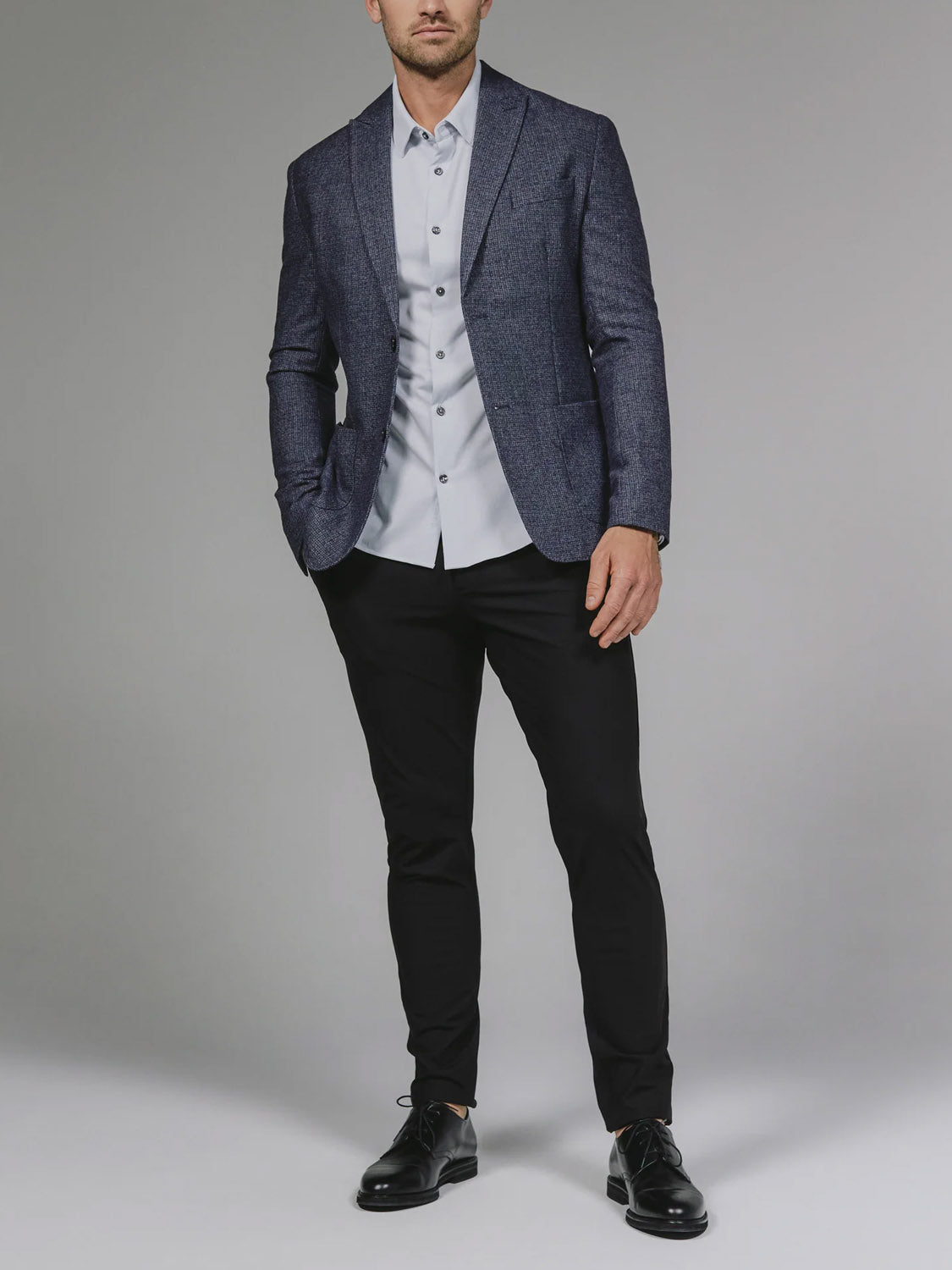 A man stands in a studio against a plain gray background wearing the 7 Diamonds Belfast Blazer over a light gray dress shirt, paired with black pants and black dress shoes, his left hand casually in his pocket.