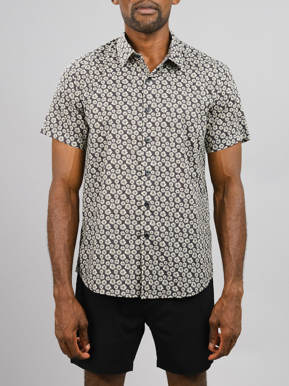 A man wearing the ID Menswear Bellis Printed Cotton Shirt with black and white floral pattern and black shorts stands against a plain light gray background.