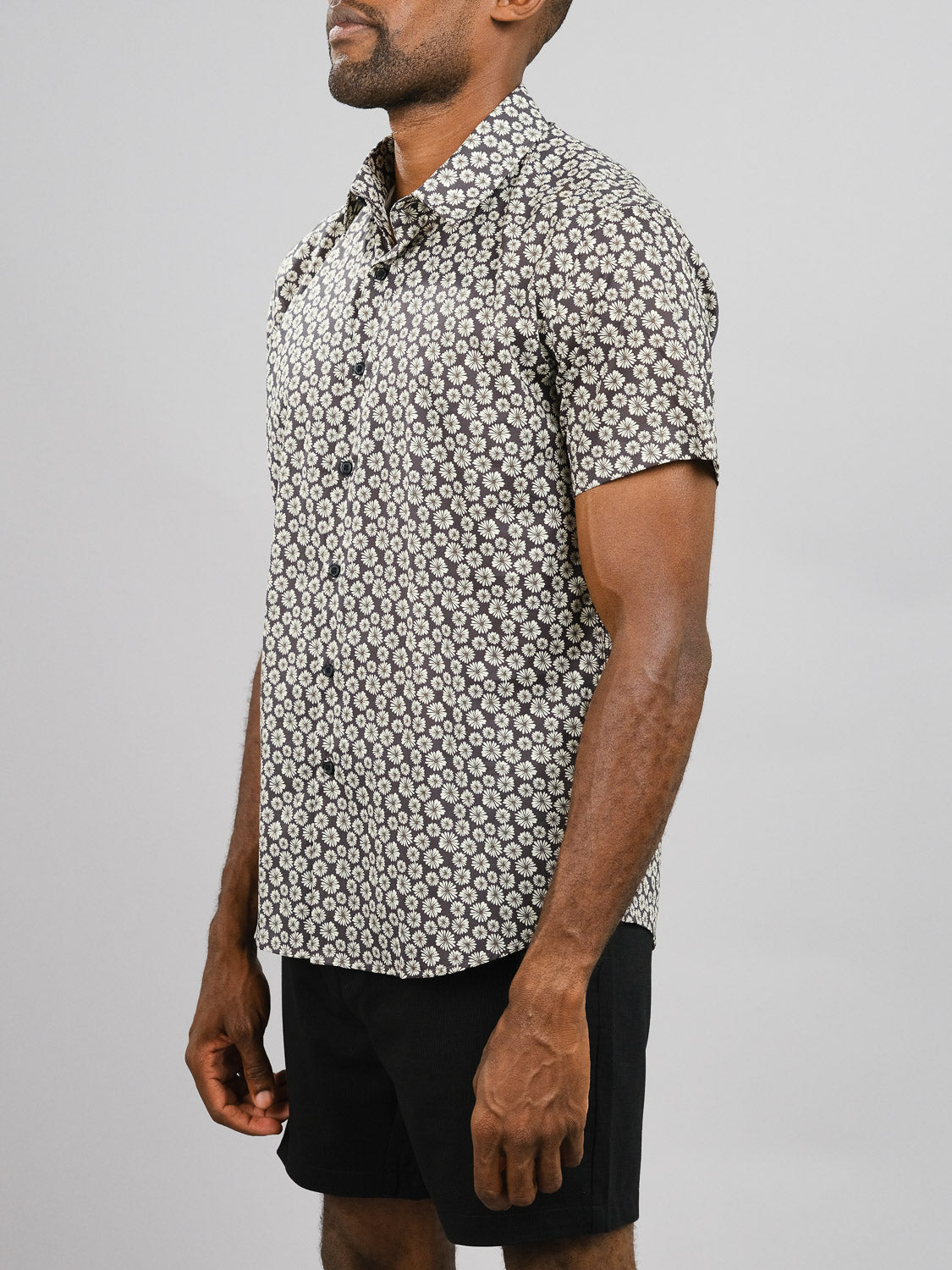 A man wears the Bellis Printed Cotton Shirt by ID Menswear, featuring a white floral print on black, paired with black shorts, standing against a plain light gray background.