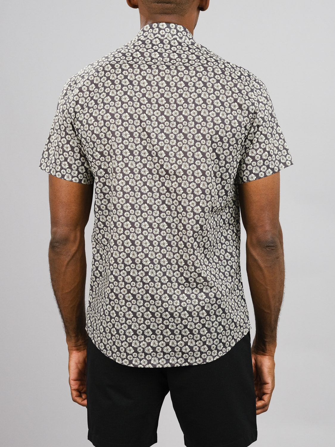 A person faces away from the camera, wearing the Bellis Printed Cotton Shirt by ID Menswear, which features a black and white circular pattern, paired with black shorts against a plain light gray background.