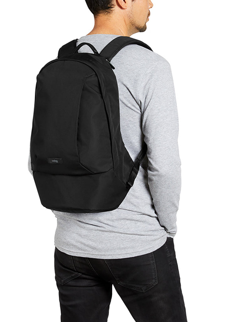 A man in a light gray long-sleeve shirt and black jeans stands with his back turned, carrying the Bellroy Classic Backpack - 20L, a sleek urban bag made from recycled materials.