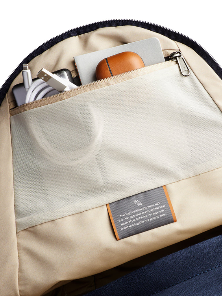 The Bellroy Classic Backpack - 20L, made from recycled, water-resistant materials, features a beige interior with a mesh pocket holding a charging cable, laptop or tablet, brown case, and zipper pouch, plus a stitched gray label inside.