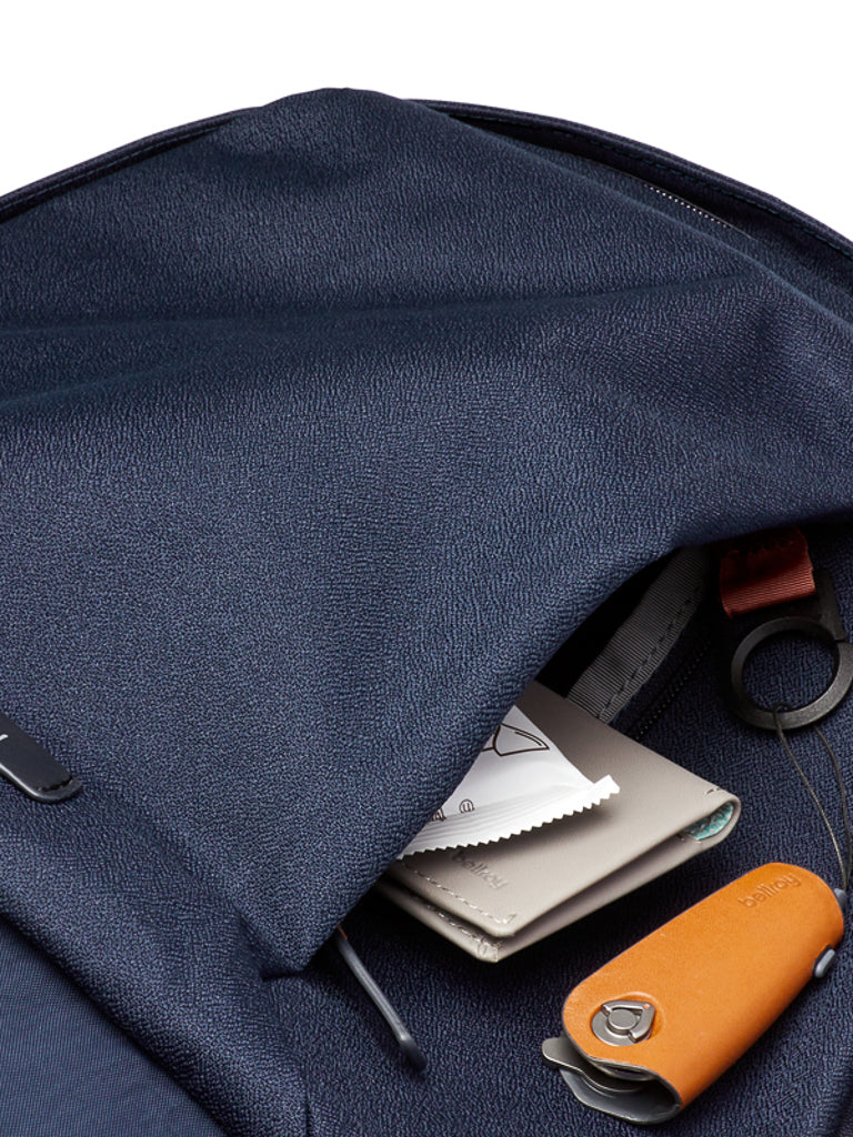 A close-up of the Bellroy Classic Backpack - 20L in navy blue shows its front pocket partially unzipped, revealing a wallet, lens cleaning cloth, circular keyring, and an orange key organizer inside.
