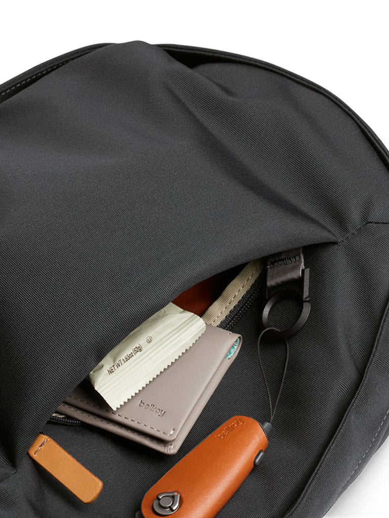 A close-up of the open front pocket of the Bellroy Classic Backpack - 20L reveals a leather wallet, a small packet, a key holder with orange and brown accents, and a fabric cleaning cloth.