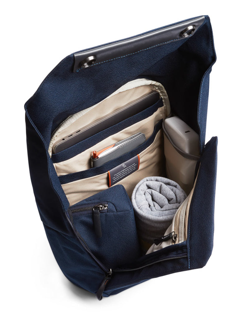 The Bellroy Melbourne Backpack Compact - 13L in navy blue is shown open, with a rolled gray towel, water bottle, zippered pouch, pen, and electronic devices neatly arranged inside its beige compartments.