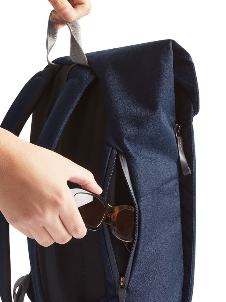 A hand places sunglasses into the front zip pocket of the Bellroy Melbourne Backpack Compact - 13L in navy blue, which stands upright by its top handle against a white background.