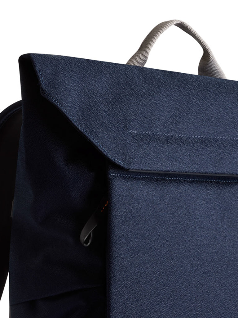 Close-up of the Bellroy Melbourne Backpack Compact (13L) in navy blue, showcasing its textured minimalist design with a front flap, gray handle, and zippered pocket—perfect for urban professionals on the move.