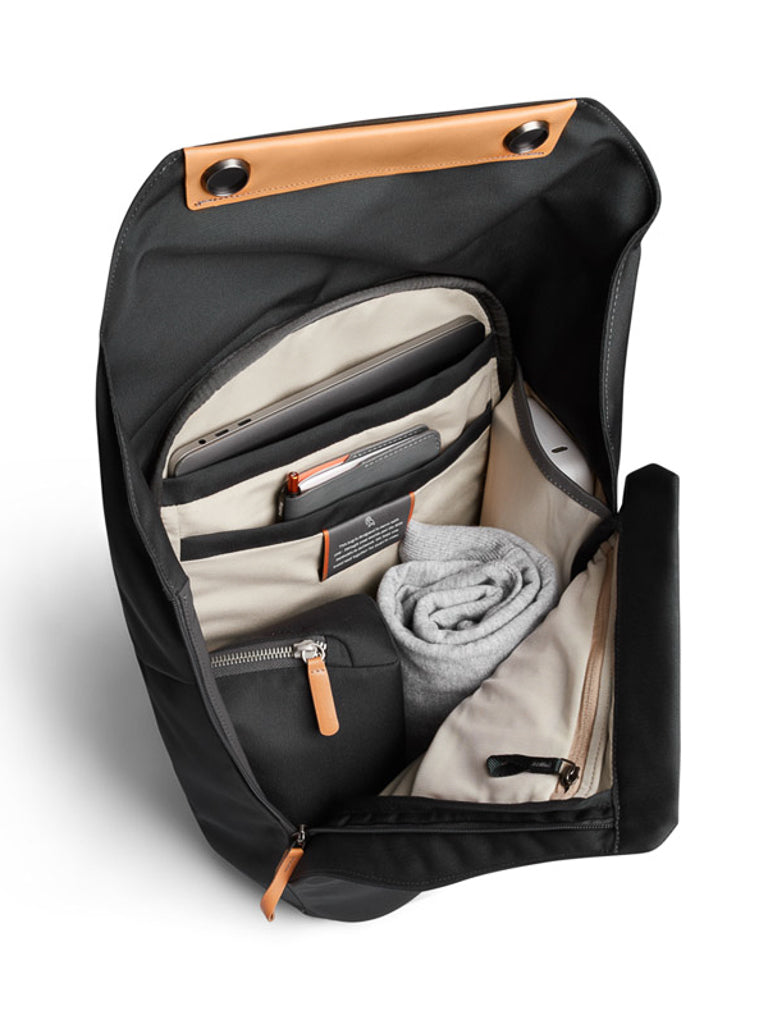 The Bellroy Melbourne Backpack Compact - 13L features an open black design with a beige-lined interior, holding a rolled grey garment, zip pouch, notebook, and pen. Brown leather accents on top and zipper pulls add a polished touch to this minimalist backpack.