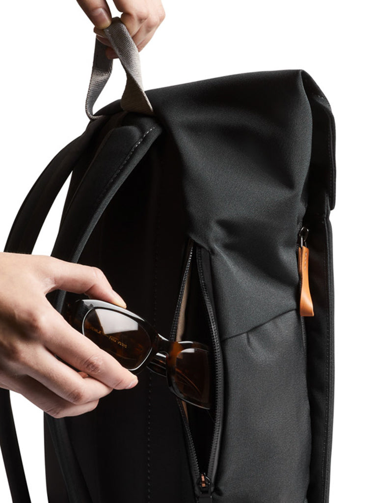 A hand places sunglasses into the zippered side pocket of the black Bellroy Melbourne Backpack Compact - 13L with a tan zipper pull, set against a white background.