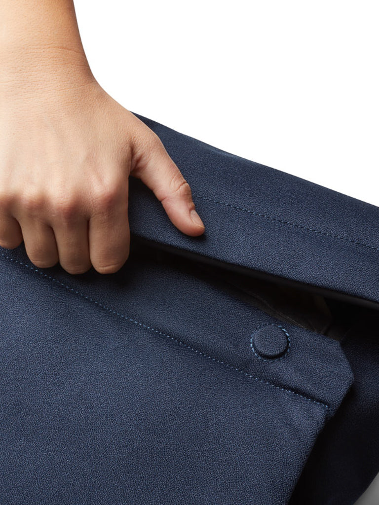 A hand lifts the flap of a Bellroy Melbourne Backpack - 18L, dark blue in color, revealing a button beneath, highlighting the clean lines and minimalist design against a plain white background.