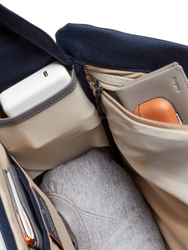 The Bellroy Melbourne Backpack - 18L opens to reveal organized compartments for a sweater, charger, sunglasses case, and beige wallet. Its beige interior contrasts with the dark blue exterior—ideal for carrying stylish urban essentials.