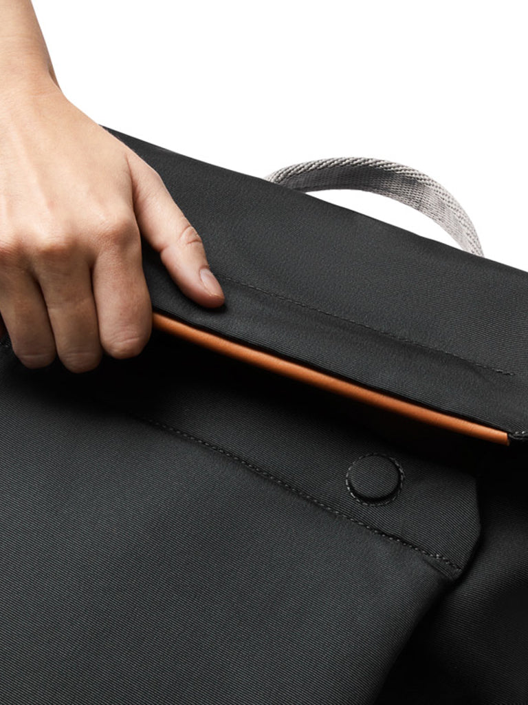 A hand places a slim, brown device into the outer pocket of the Bellroy Melbourne Backpack - 18L, a minimalist black fabric bag featuring a button closure and white strap.