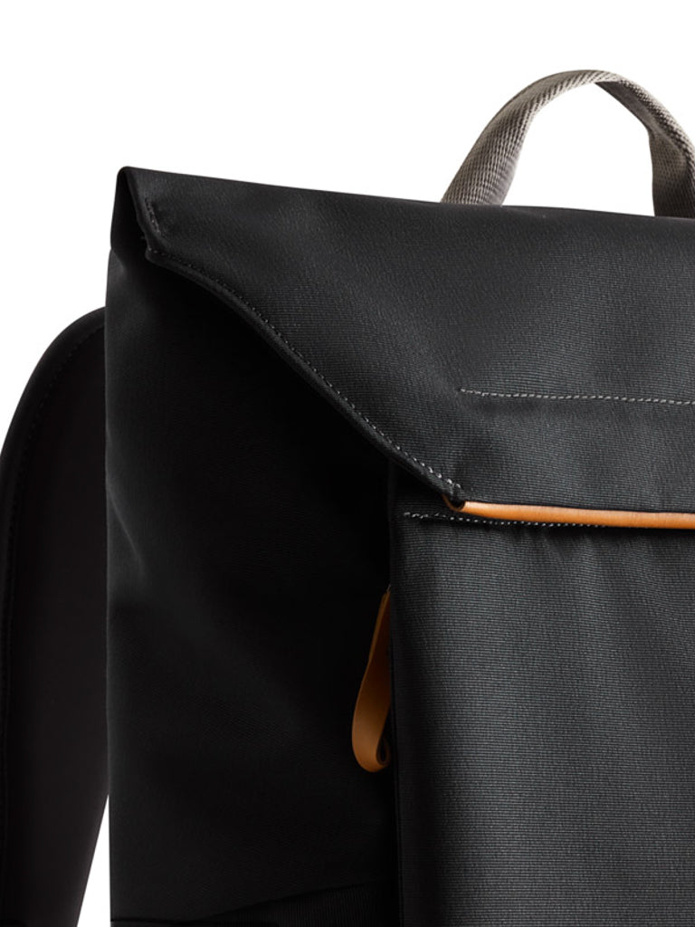 Close-up of the Bellroy Melbourne Backpack - 18L in black, featuring a minimalist design with a flap closure, beige handle, and brown accent near the top, shown against a white background.