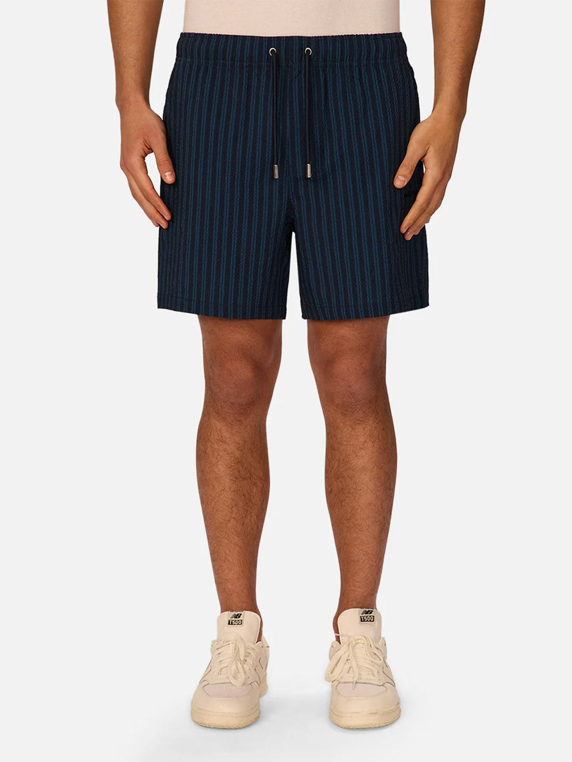 Someone wearing Industrie’s Berkeley Swim Short—navy blue pinstriped seersucker with a drawstring waist—and light sneakers, stands against a plain light background. Only their lower half is visible.