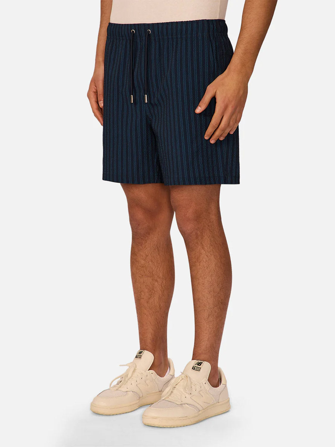 Only the lower half of a person is visible, wearing Industrie’s Berkeley Swim Short in navy seersucker with a drawstring, paired with off-white sneakers and a light beige t-shirt against a plain light background.