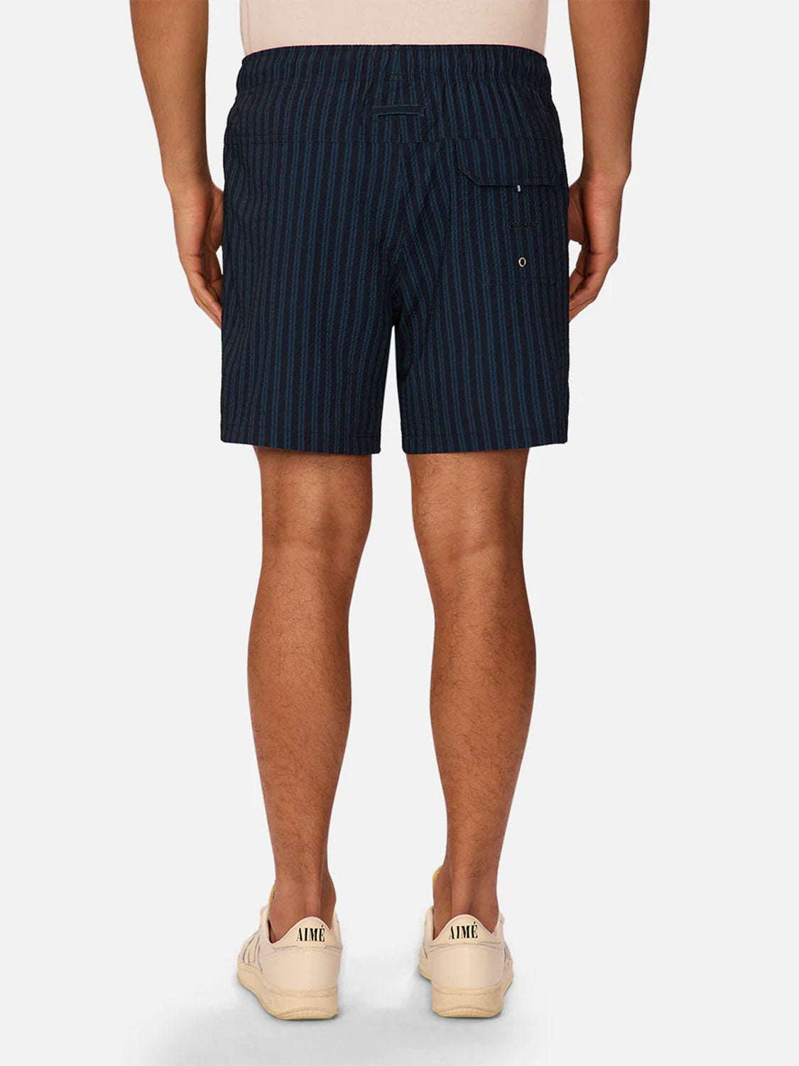 A person stands facing away in Industrie’s Berkeley Swim Short—navy pinstriped seersucker, with a buttoned back pocket and metal grommet—paired with a light beige shirt and beige sneakers, set against a plain white background.