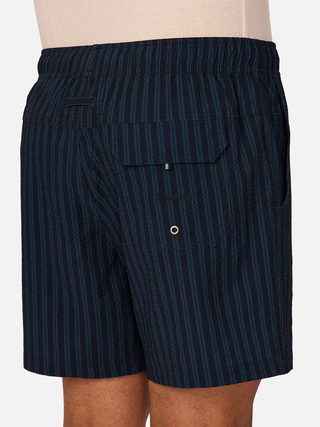 A person wears Industrie’s Berkeley Swim Short—dark navy with vertical teal stripes. These quick-dry shorts have an elastic waistband and a back flap pocket with button and grommet.