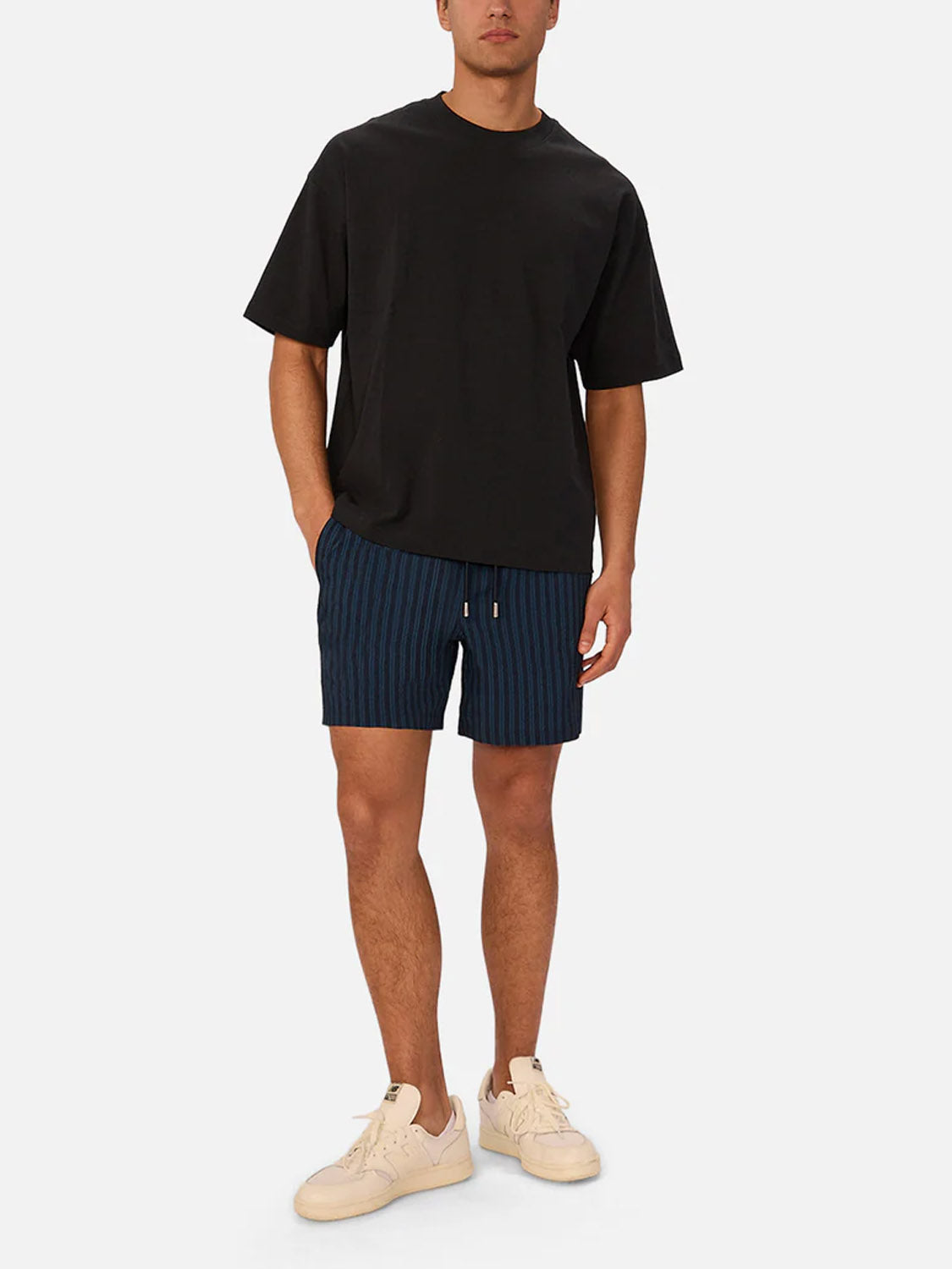 A man stands against a plain light background wearing Industrie’s Berkeley Swim Short in navy seersucker, paired with a loose black t-shirt and off-white sneakers.