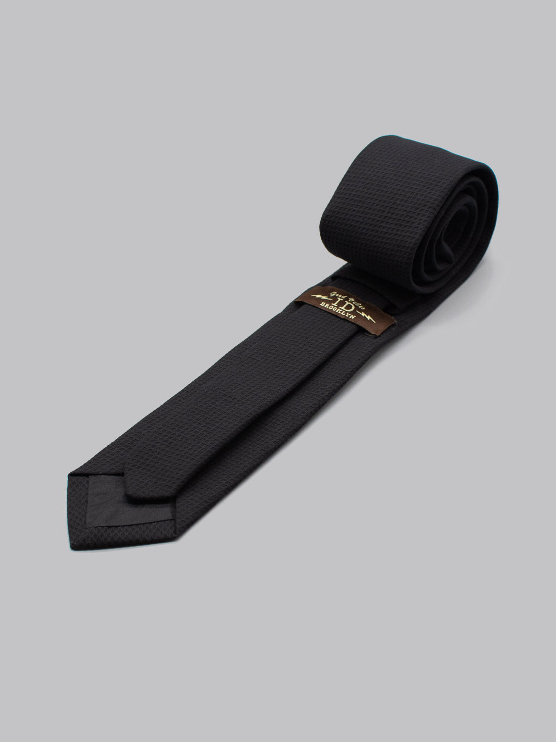 The ID Cotton Tie Berlin by ID Menswear is a black cotton tie with a subtle textured pattern, neatly rolled on a light gray background. A brown and gold brand label inside adds refined elegance to your business formal look.