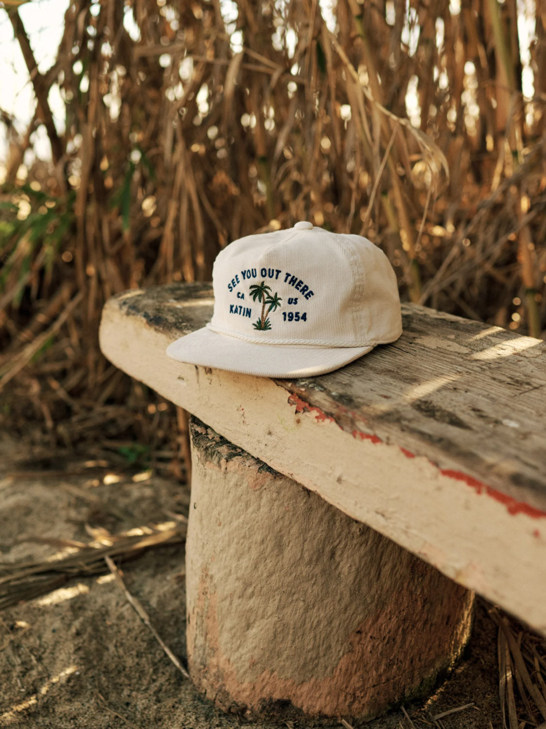 The Katin Bermuda Hat, a beige 5-panel cotton corduroy hat with palm tree embroidery and See You Out There Katin 1954 text, sits on a weathered wooden bench amidst tall grass.