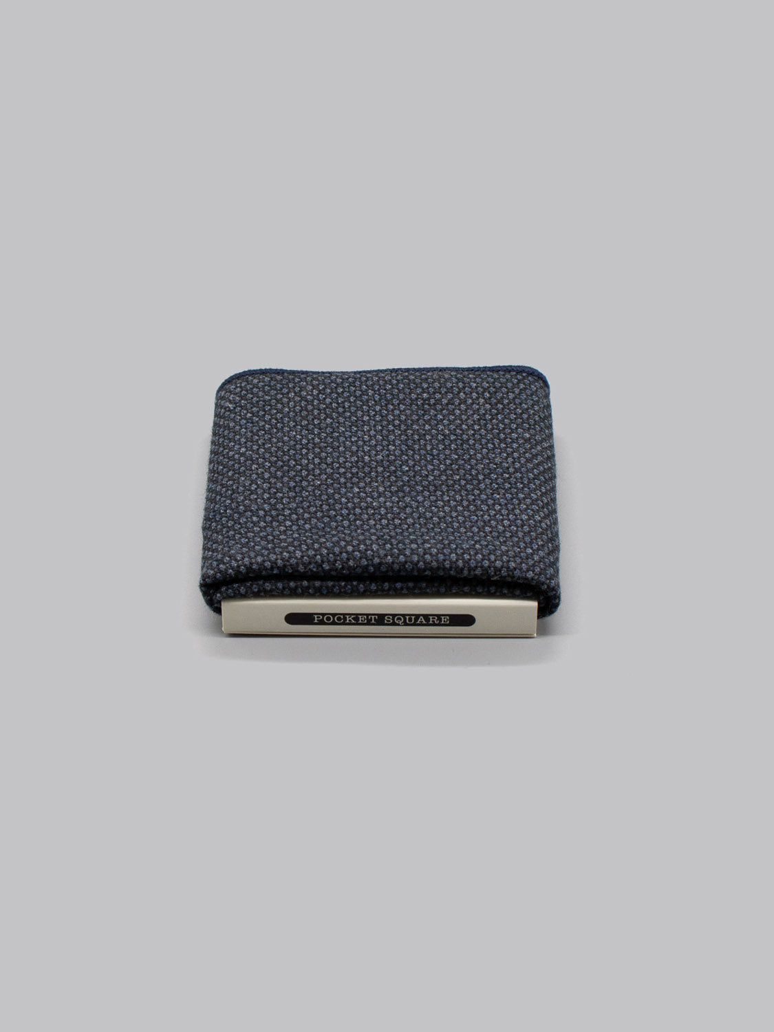The Bern Pocket Square by ID Menswear, handmade in Brooklyn from 100% cotton with a subtle dotted pattern, is folded and secured by a silver POCKET SQUARE clip against a plain light gray background.