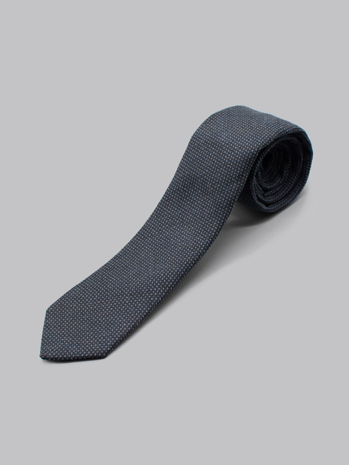 The ID Menswear ID Cotton Tie Bern, a dark blue cotton tie with a subtle dotted pattern, is handmade in Brooklyn and neatly rolled on a plain light gray background.