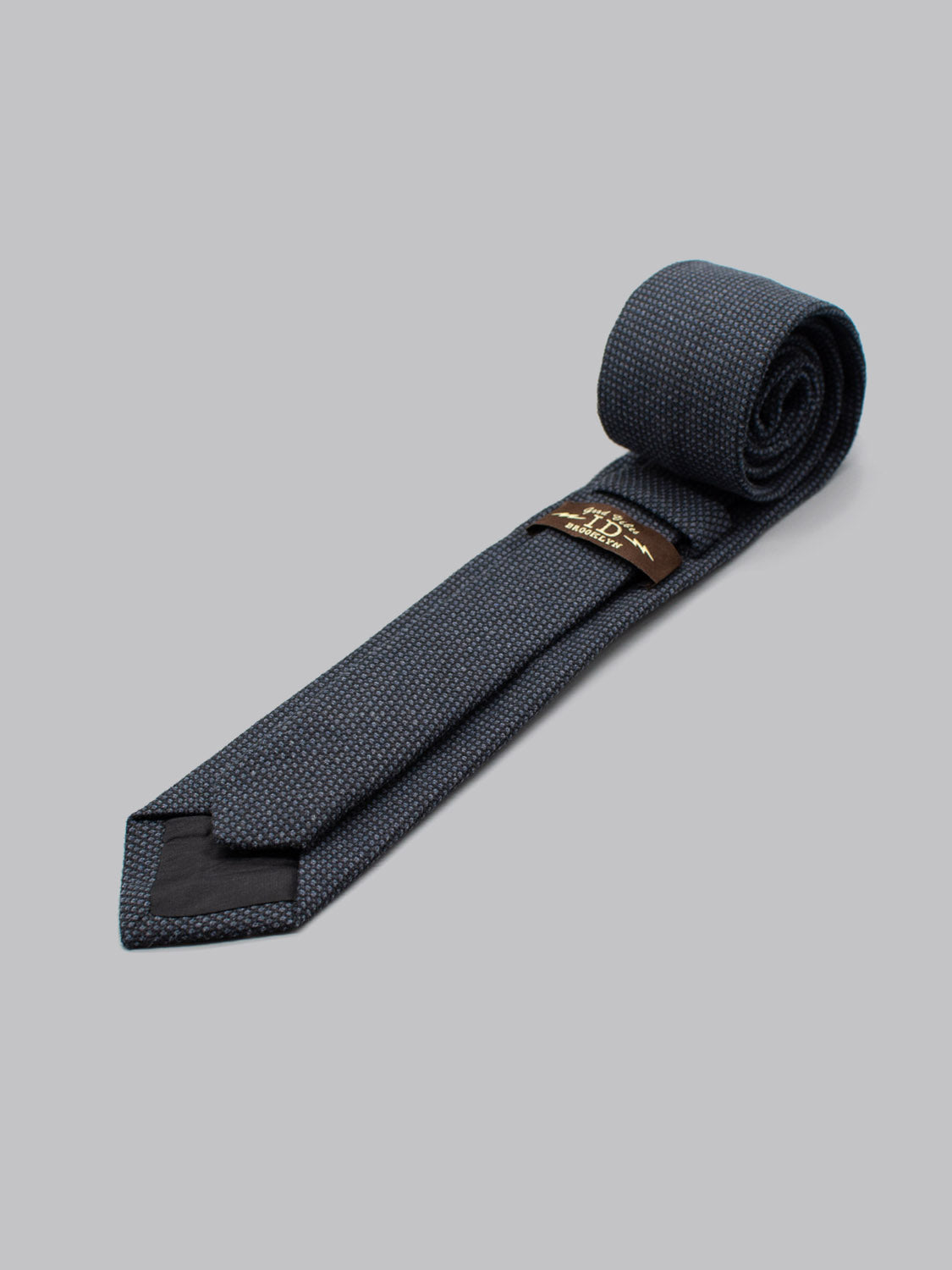 The ID Menswear ID Cotton Tie Bern, a dark blue textured cotton tie, is handmade in Brooklyn and neatly rolled on a light gray background. A small brown and gold label appears near the wider end of this business formal accessory.