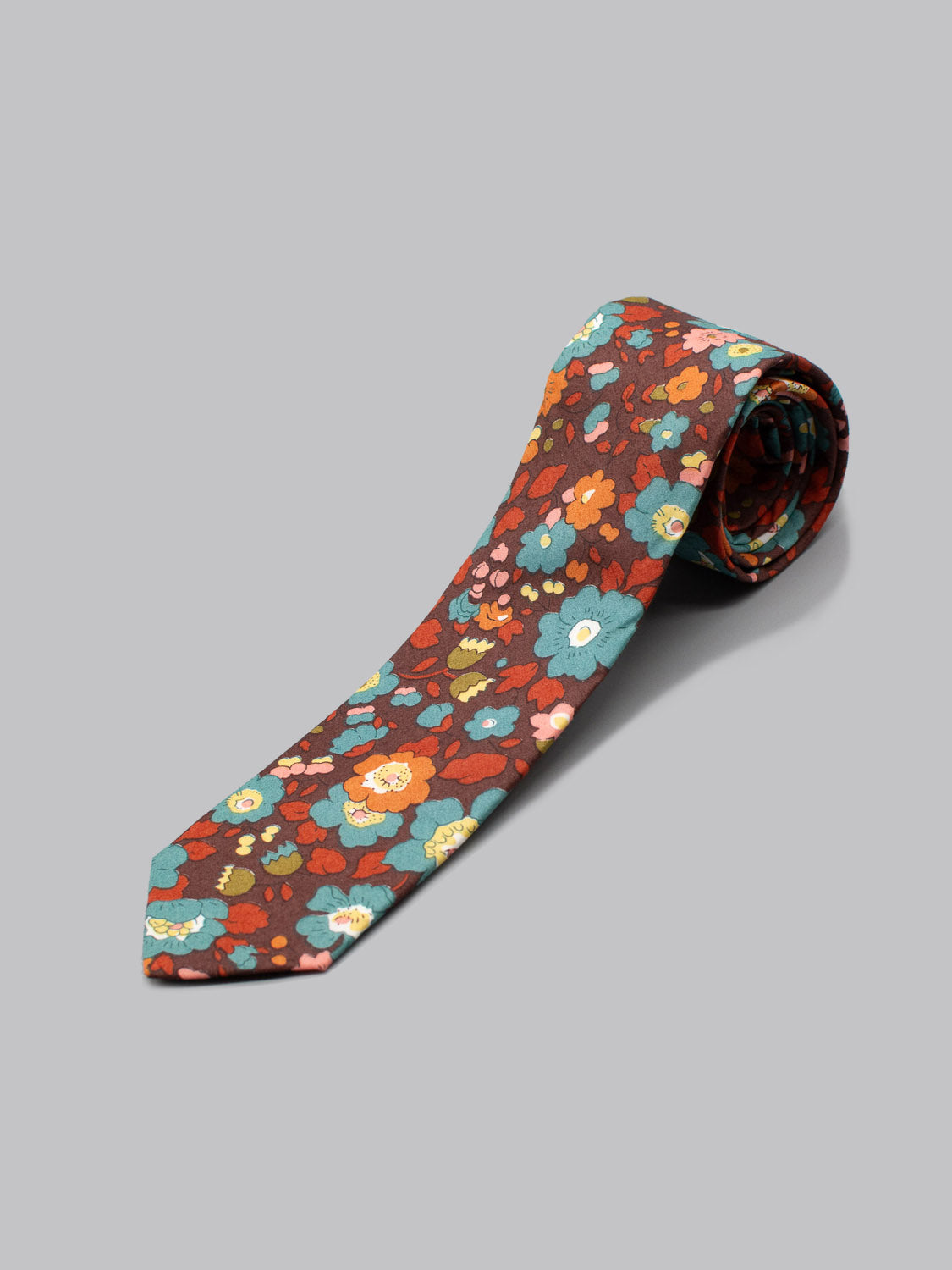 The Liberty of London Tana Lawn™ Betsy Tie by ID Menswear features a vibrant blue, orange, yellow, and pink floral print on rich brown, handmade and shown rolled up on a light gray surface.