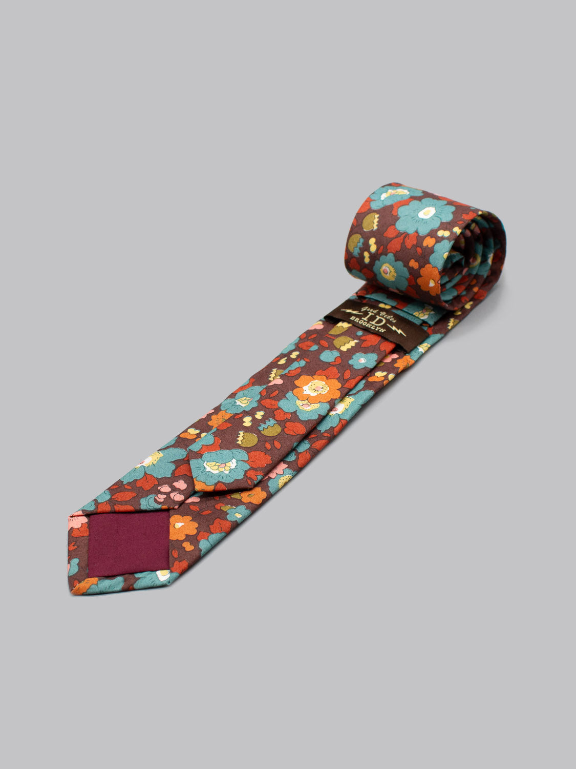 The Liberty of London Tana Lawn™ Betsy Tie by ID Menswear features a red background with blue, orange, and yellow floral patterns, handmade from premium fabric for a silk-like feel and shown rolled up on a light gray surface.